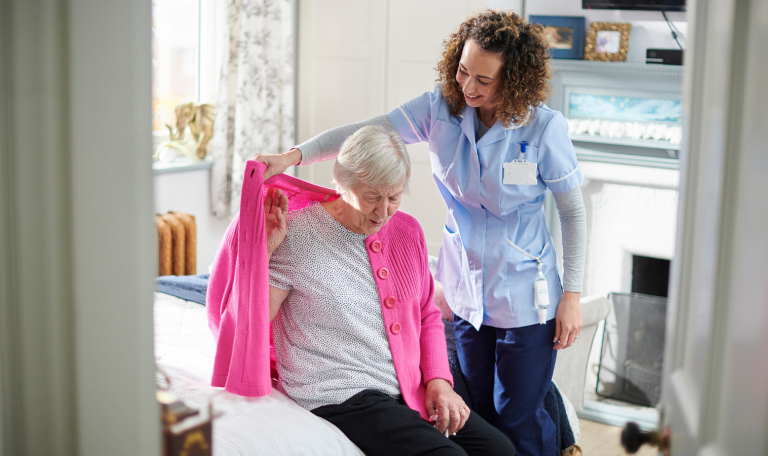 An older woman sat on a bed being helped by a carer to put a cardigan on 