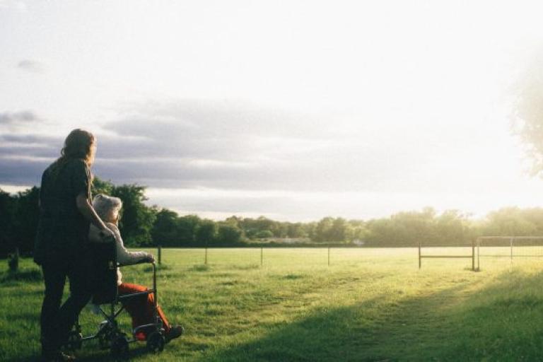 A young woman pushes an older person in a wheelchair in a field where the sun is setting