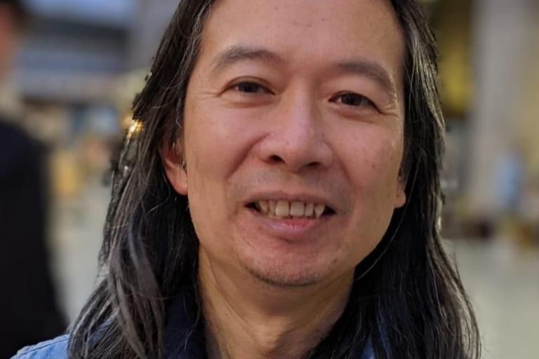head and shoulders shot of Andrew Chiu, he is an asian man and has long hair and is wearing a blue top, he's smiling at the camera